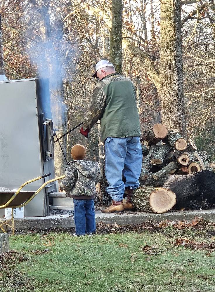 An operator and child tending an outdoor wood stove beside cut logs